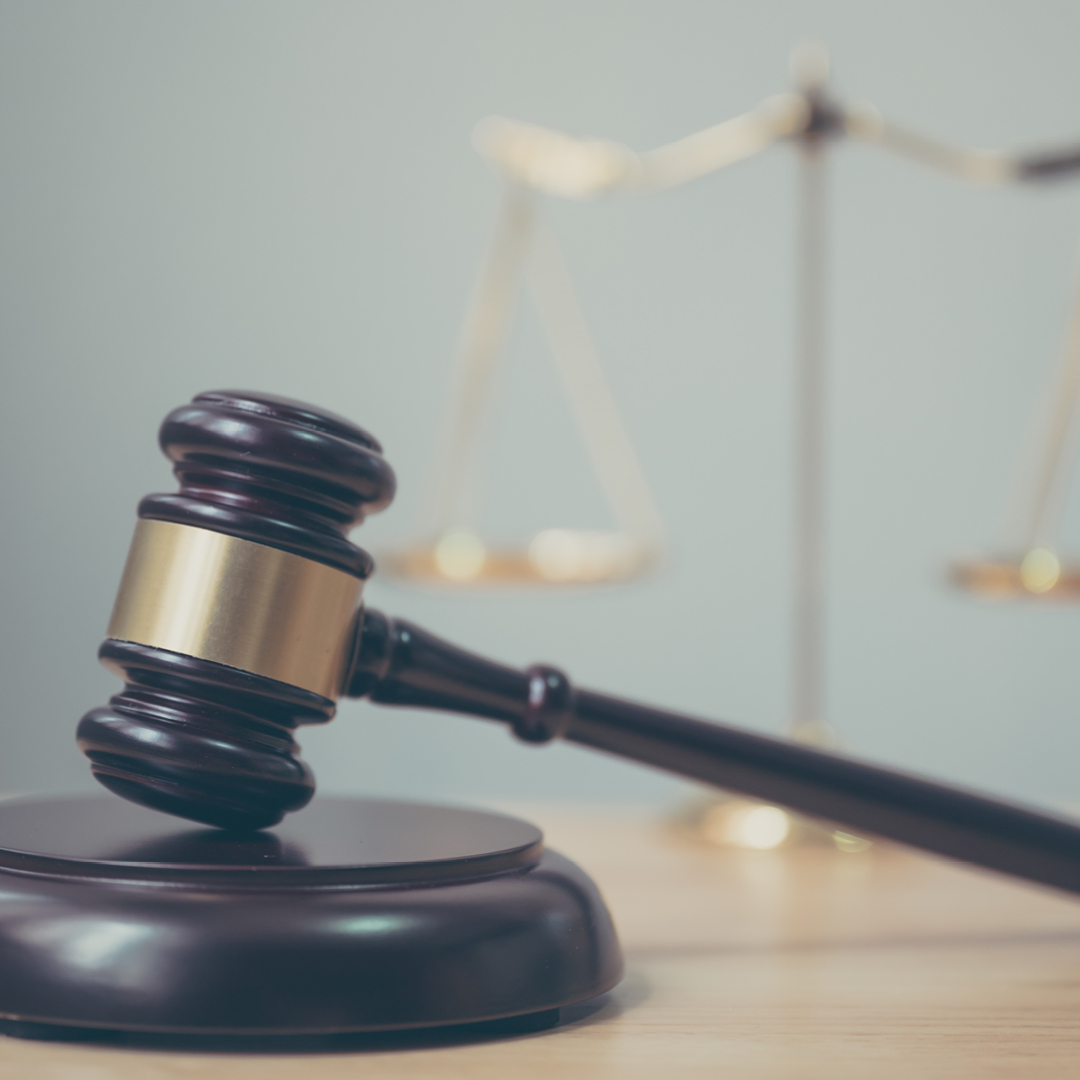 A wooden judge's gavel and sounding block rest on a light wood surface, with a blurry gold scale of justice in the background.