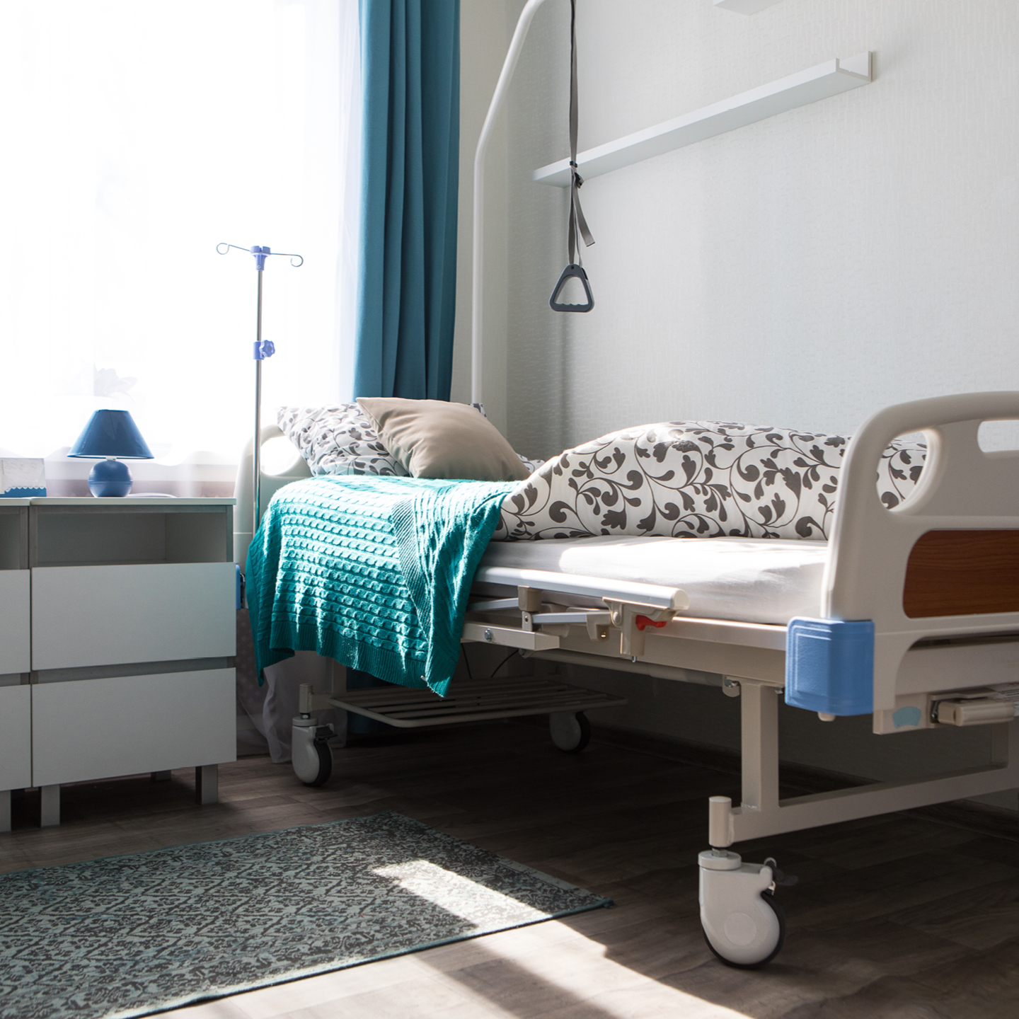 An adjustable hospital bed with patterned bedding, a teal blanket, a pillow, an overhead grab bar, and an IV pole stands by a window with teal curtains, next to a white bedside table with a blue lamp on a patterned rug.