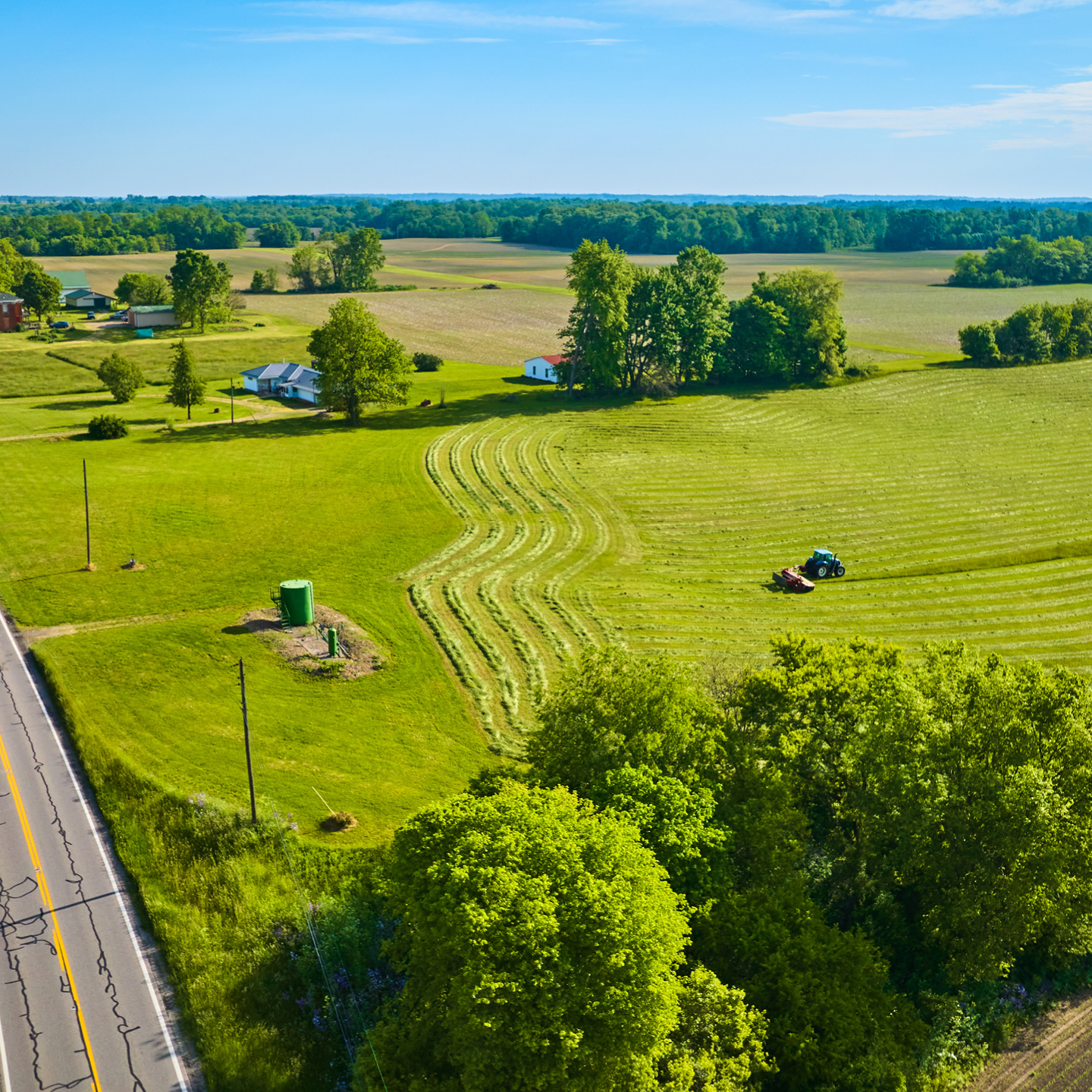 An aerial view of a vibrant green agricultural landscape under a clear blue sky, featuring a blue tractor operating in a large, open field.