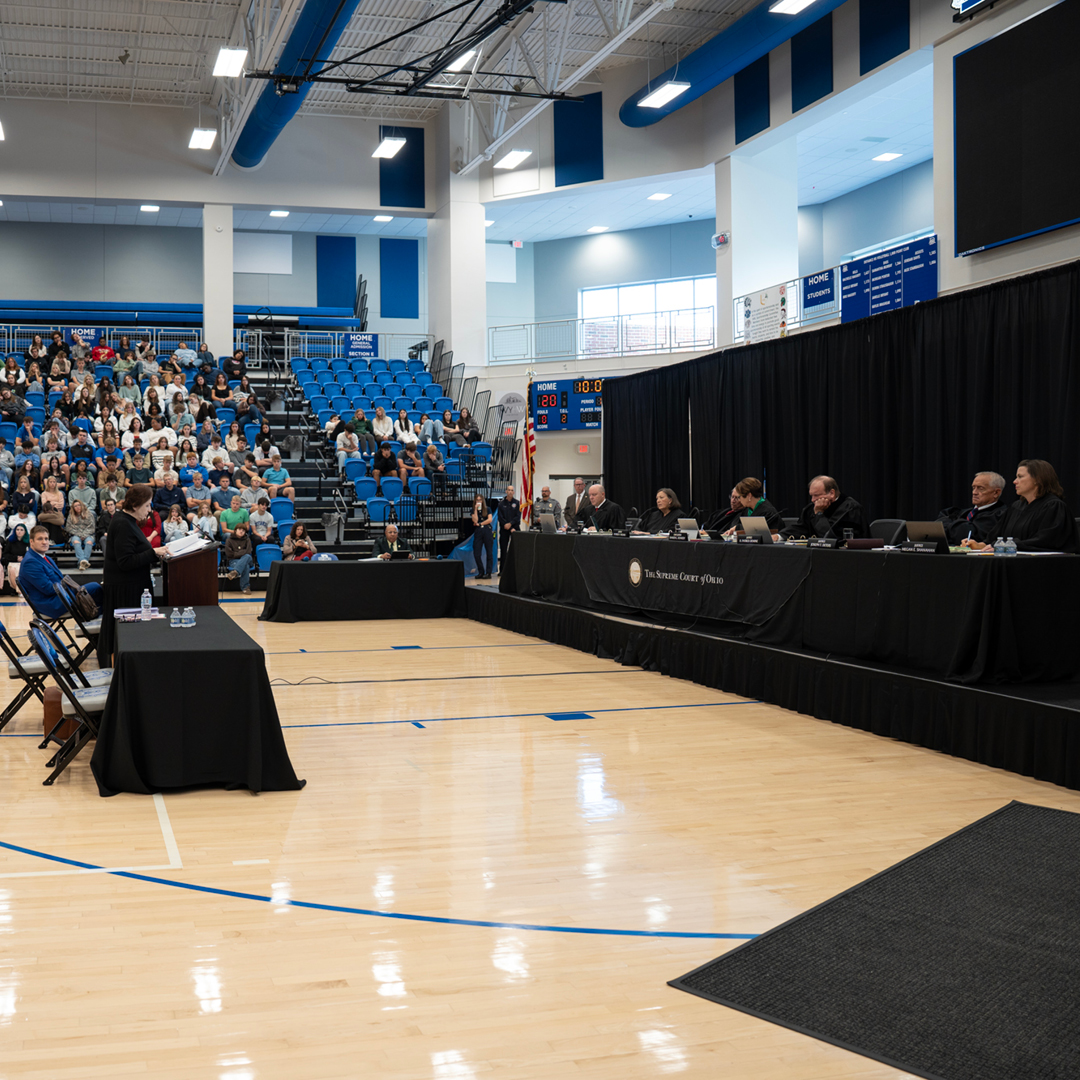 A gym with the stands full at the front of the room is a table with seven justices sitting wearing black judicial robes