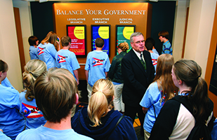 Image of Visitor Education Center longtime volunteer Pete Miller speaking to a group of students in front of the Balance Your Government interactive display