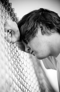 Image of a young man resting his head on his forearm, leaning against a chain link fence