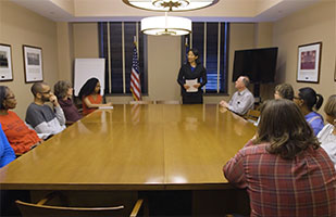 Image of a group of people sitting at a conference table listening to a woman standing at the head of the table
