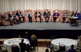 Image of a group of men and women sitting in armchairs on a stage speaking to an audience of people sitting at round tables