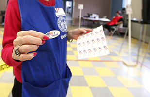 Image of a female poll worker wearing a blue apron and holding out an I voted sticker