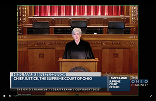 Image is of a screenshot of Chief Justice O'Connor speaking at a podium in front of the Supreme Court bench.