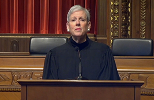Image of Chief Justice O'Connor in her black judicial robe, standing at a podium in front of the Supreme Court Courtroom bench