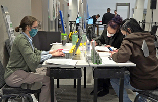 Image of 3 women, all wearing masks, sitting and working at a table, with 3 others in the bacground