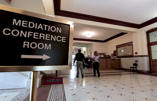 Image of two people in a building hallway, with a sign in the foreground that says Mediation Conference Room and an arrow pointing right