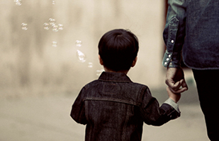 Image of the back of a young boy with dark hair, wearing a denim jacket and holding the hand of an adult