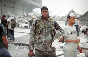Image of Jason Thomas in  his military fatigues looking at the camera while standing among the rubble of the World Trade Center, with several other people in the background