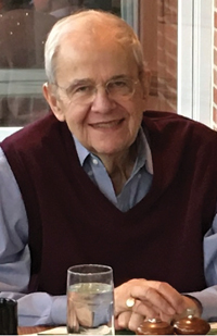 Image of a man wearing a sweater vest sitting at a table smiling with his hands folded in front of him