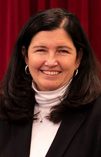 Image of a headshot of Lyn Tolan in front of a red curtain wearing a black suit jacket and white turtleneck sweater