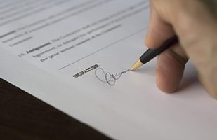 Image shows part of a white paper form and a hand appearing to write a person's signature on the form