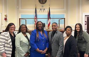 Image of five women and one man standing side-by-side in front of a judge's bench in a courtroom