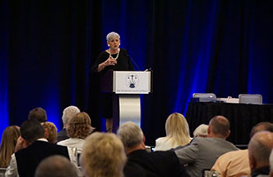 Woman on a podium with a blue backdrop speaking to an audience.