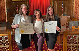 Three Caucasian women standing next to each other. The mother stands between her two daughters holding their law certificates in a courtroom.