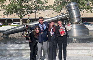 Image of two boys and two girls, all wearing suits, wearing medals, posing in front of the giant, stainless steel gavel in the south reflecting pool of the Thomas J. Moyer Ohio Judicial Center. One boy is holding a trophy, while a girl is holding a plaque and certificate.