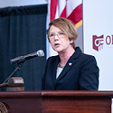 Woman wearing a blue suit speaking from a wooden podium to a room full of men and women.