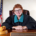 Image of a female judge with red hair and wearing a black judicial robe seated at a wooden table with a wooden gavel at her side.