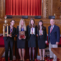 Image of five girls, all wearing blue suits and medals around their neck, standing next to a man, also wearing a blue suit, in the courtroom of the Thomas J. Moyer Ohio Judicial Center. The first girl on the left is holding a large trophy and the second girl on the left is holding a plaque.