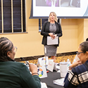 A woman, wearing a grey and black pattern dress and a black jacket, speaks to a room full of men and women.