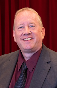 Image of a man wearing a dark grey suit, maroon shirt, and blue tie.