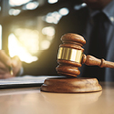 Image of a wooden gavel resting on a desk. In the background, a man writes on some documents.