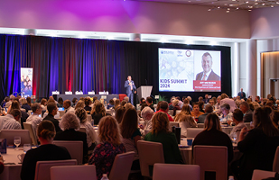 Image of a man speaking from a podium to a large room full of men and women seated and round tables.