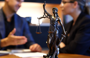 Image of a small statue of Lady Justice with a man and woman talking in the background.