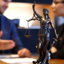 Image of a small statue of Lady Justice with a man and woman talking in the background.