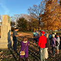 A man wearing a grey short-sleeve shirt stands in front of a large monument reading from a paper to a group of men, women and children.