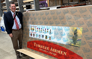 A man wearing brown dress pants, white dress shirt, red tie, and blue jacket stands next to a large, rectangular board displaying images of various military medals and a red sign that says: 'Tuskegee Airmen. Portraits in Courage.'