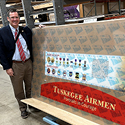 A man wearing brown dress pants, white dress shirt, red tie, and blue jacket stands next to a large, rectangular board displaying images of various military medals and a red sign that says: 'Tuskegee Airmen. Portraits in Courage.'