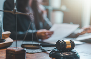 Image of a wooden gavel and scales of justice sitting on a wooden desk. In the background, a person holds a document.