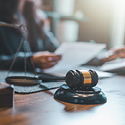 Image of a wooden gavel and scales of justice sitting on a wooden desk. In the background, a person holds a document.