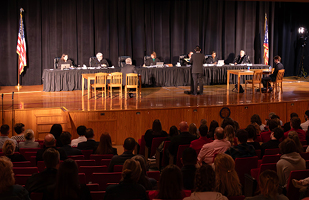 Image of a man wearing a black suit standing at a podium facing a long table with black skirting. Seated at the long table are 6 men and women wearing black judicial robes. All are on a stage in an auditorium full of people.