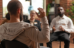 Image of a man with a beard and short-cropped hair wearing a grey hooded sweatshirt. He has his right arm raised and is seated in a group circle.