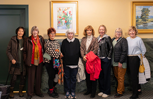 Eight women standing side-by-side in front of a wall with a painting of flowers and a painting of a pond lined by trees.