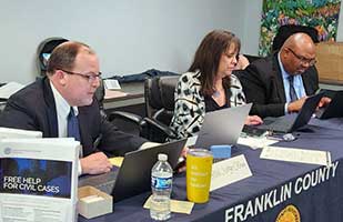 Two men and a woman seated at a long table with a blue table cloth that says 'Franklin County Municipal Court'. All three people are using laptops.