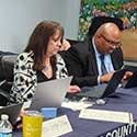 A woman and a man seated at a long table using laptops.