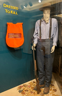 Image of a manequin wearing dark pants, a blue shirt and suspenders next to a bright orange vest.