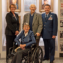 A woman wearing a blue suit stands next to a man wearing a grey blazer and another man wearing an Air Force dress uniform. All are standing behind a woman seated in a wheelchair.