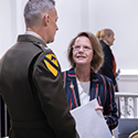 A woman wearing a red, white, and blue stiped blazer speaks with a man wearing a military dress uniform.