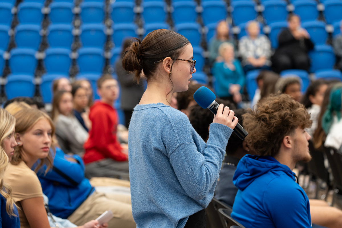 A teenager speaking into a microphone surrounded by her peers