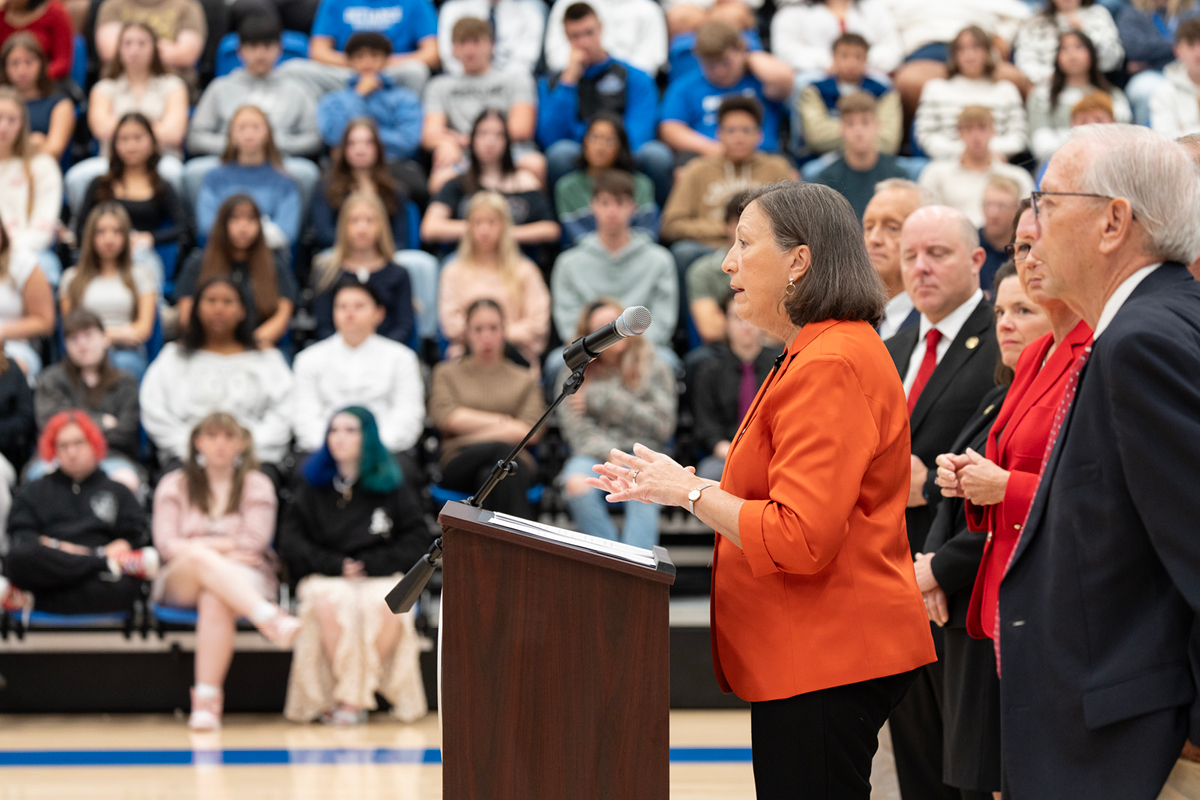 A woman at a podium speaking to a large crowd in a gym 