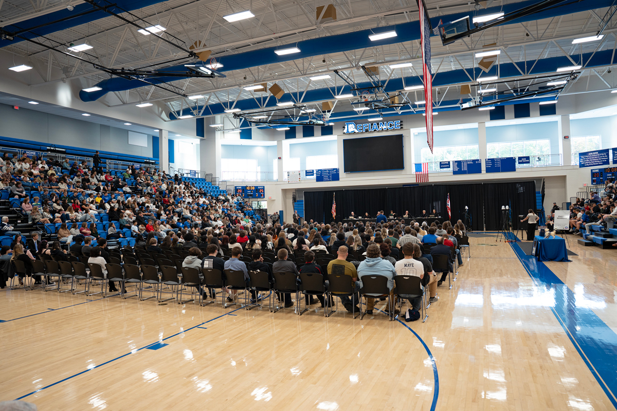 A gym full of highschool students an a pannel of seven adults on a stage