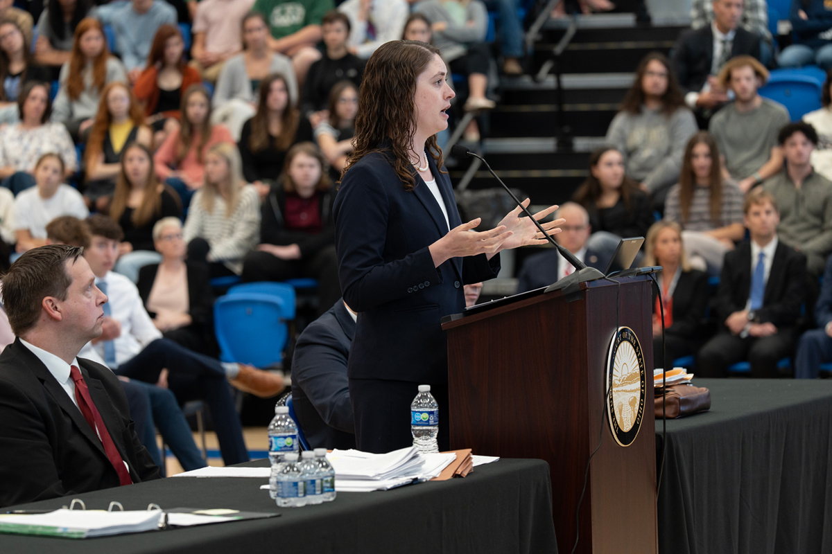 A woman speaking at a smaller podium surrounded by tables with papers and a seated man