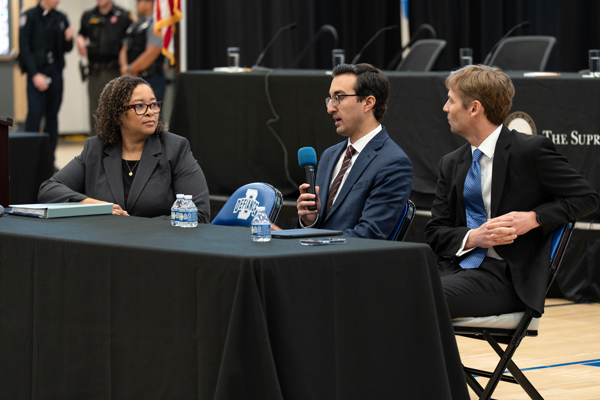 Three adults seated at a table speaking to a gym full of highschool students