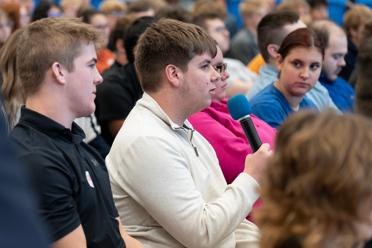 A highschool boy seated speaking into a microphone surrounded by his peers